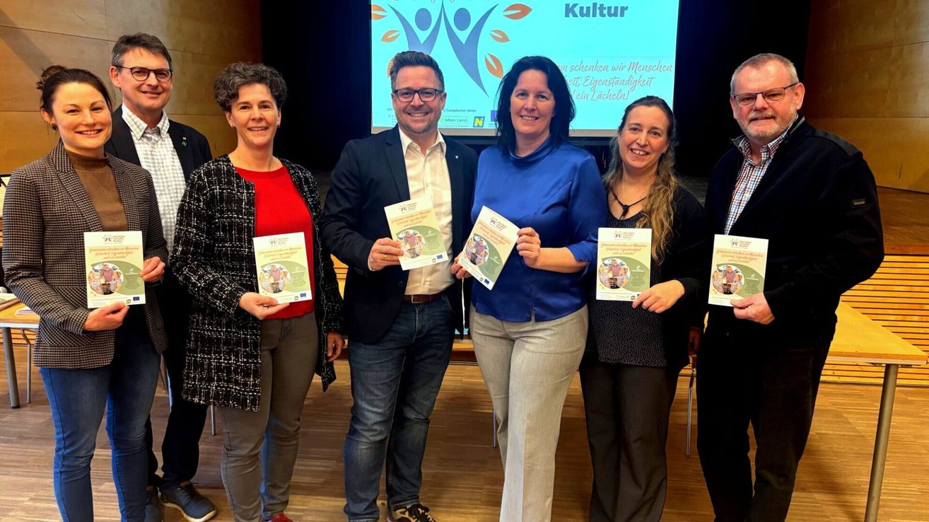 Fünf Menschen stehen zusammen auf einem Holzboden und lächeln. Sie halten ein Buch in den Händen und scheinen für ein Foto zu posieren.