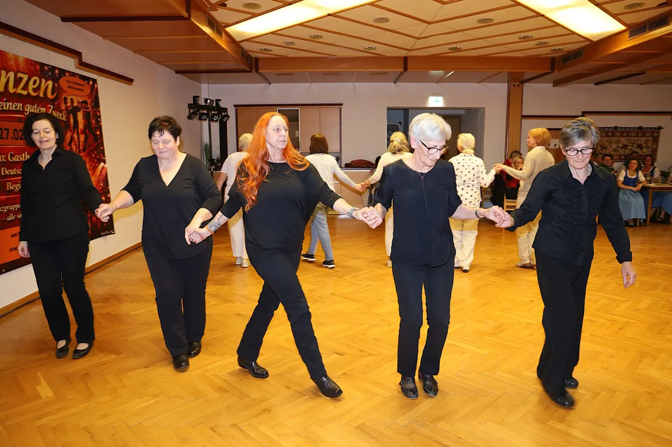 Several women in black clothes are dancing together in a room with wooden flooring. The woman in the middle has red hair and is holding hands with two other women.