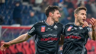 Two soccer players in black uniforms with 'Dormero Hotels' on their jerseys celebrate on a stadium field.