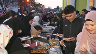 A group of people are gathered around a long table filled with various dishes, serving themselves. Some wear headscarves, and a young man holds a plate with a fork. In the background, houses and trees are visible.