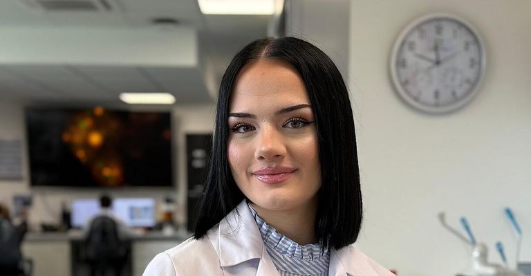Eine Frau mit kurzen schwarzen Haaren lächelt für ein Foto in einem Labor. Sie trägt einen weißen Laborkittel und ein gestreiftes Hemd. Hinter ihr ist eine Uhr an der Wand montiert.