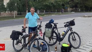 A man stands next to two bicycles, smiling. One bike has a flag, and all bikes are equipped with bags and helmets. The setting includes trees, a street, and a building in the background.