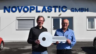 Two men stand in front of a building labeled 'OVOPLASTIC GM', holding a large white key. They are smiling and appear to be posing for a photograph.