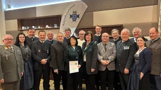 A group of adults in formal attire stand in front of a banner. A woman in the center holds a certificate, while a man to the right smiles. Behind them is a shelf with items.