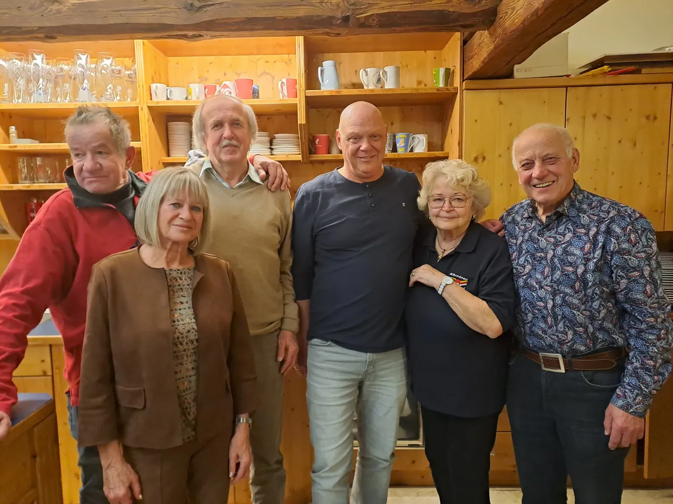 A group of older adults stands in a kitchen, smiling for a photo. The woman in the middle wears glasses. Behind them are wooden shelves with cups and plates.