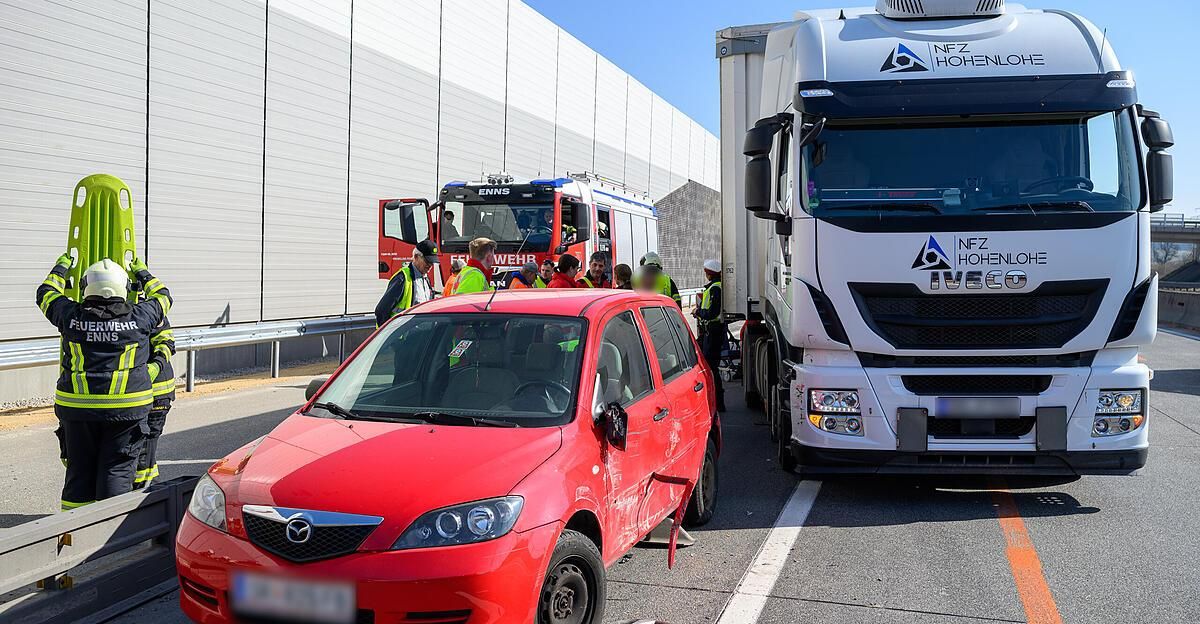 A red car is damaged on the side, with a fire truck and a truck parked nearby. Emergency personnel are attending to the scene.