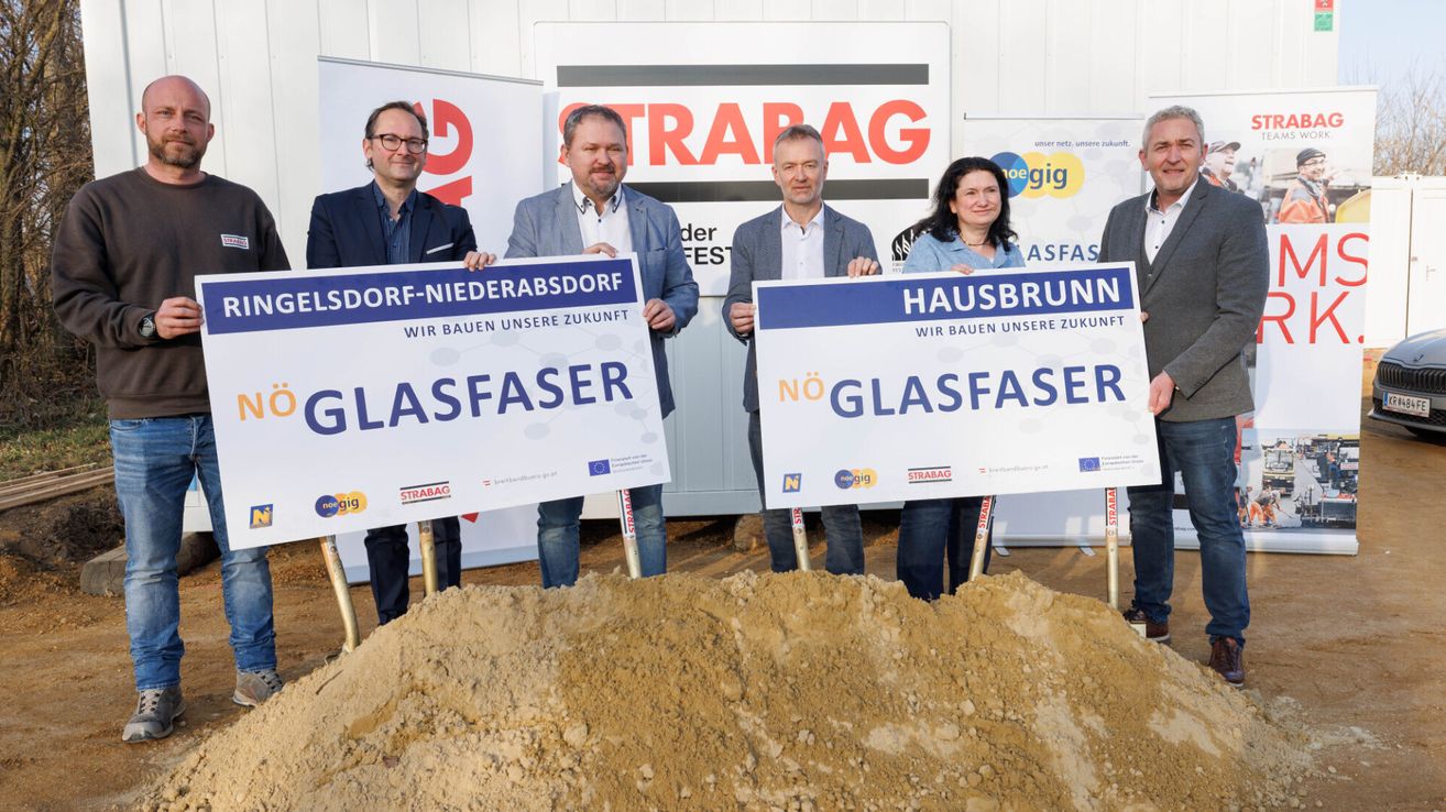 Five people stand in front of a pile of dirt. They hold signs, with one reading 'GLASFAESER' and another 'HAUSBRUNN'. Behind them, there is a banner with the word 'STRA BAG'.