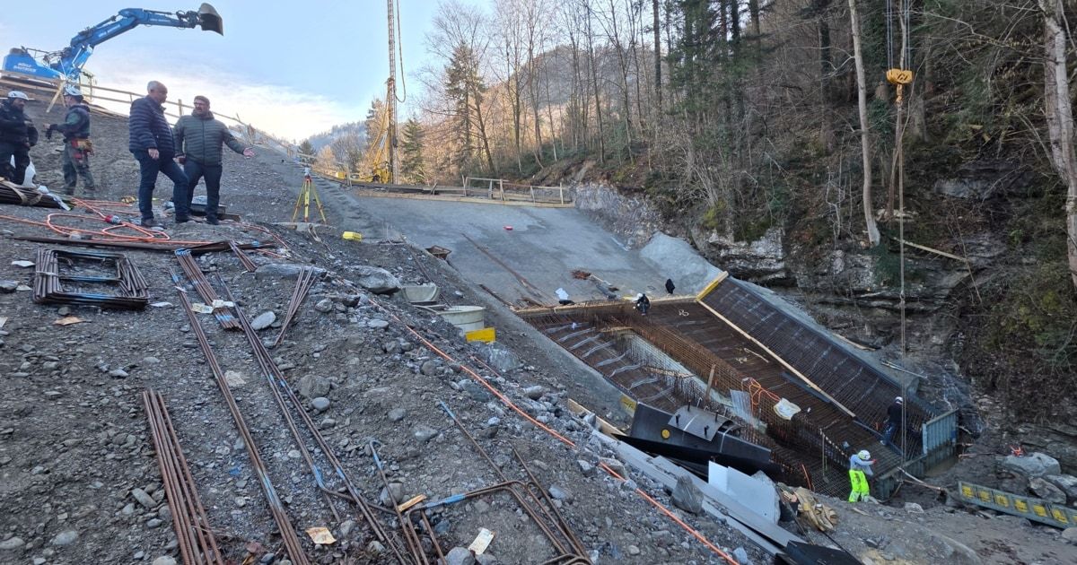 A construction site with workers in a mountainous area, featuring a bridge under construction, a crane, and scattered materials.