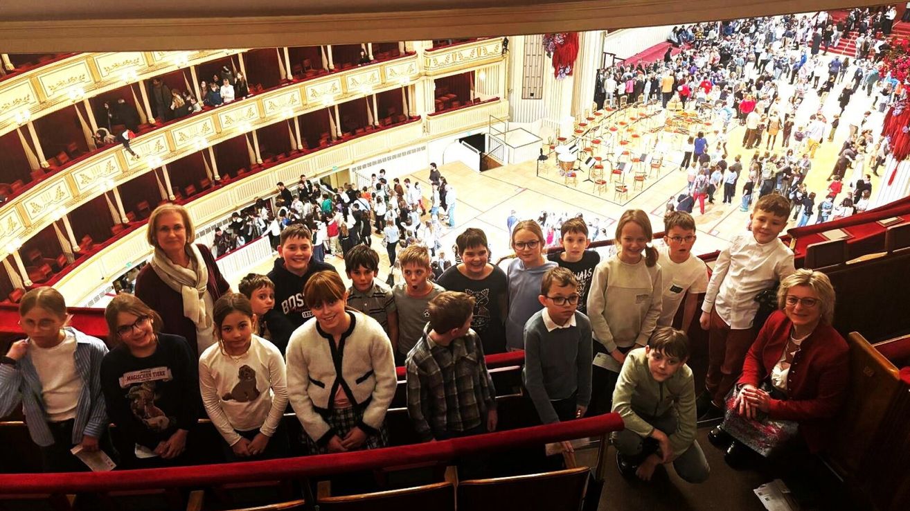 Eine Gruppe von Kindern steht in einem Auditorium mit einem großen Publikum hinter ihnen. Die Kinder tragen verschiedene Kleidungsstücke und einige haben Brillen. Das Publikum sitzt auf roten Stühlen.