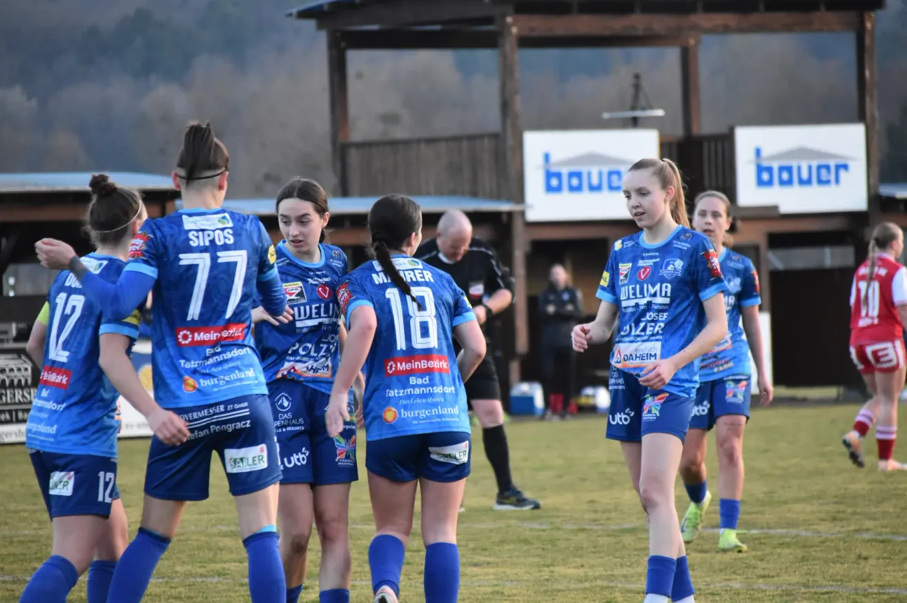 A group of young female soccer players wearing blue uniforms with numbers 77, 18, and others are standing on a field. Behind them is a man in a black shirt. There is a sign with the word 'bouc'.