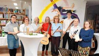A group of people posing for a photo in a room with musical instruments. A woman holds a guitar, while the others hold various woodwind instruments. A table with plants is in the foreground.