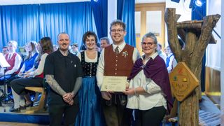 A group of people stand together smiling and posing for a photo. The man in the middle holds a certificate. The man on the left has tattoos on his arms. The woman in the middle wears a necklace. Behind them are blue curtains and people.