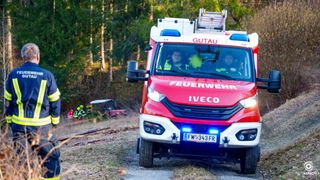 A red Iveco fire truck with two firefighters inside drives on a dirt road, with a firefighter standing on the left. Trees and a red tractor are in the background.