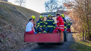 A group of firefighters wearing yellow and blue uniforms, along with a woman in a white shirt, ride in the back of a red tractor. They are on a road, possibly heading to a fire.