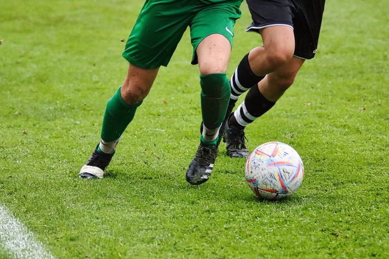 Two soccer players are on a field. One is wearing green and the other black. They are kicking a ball.