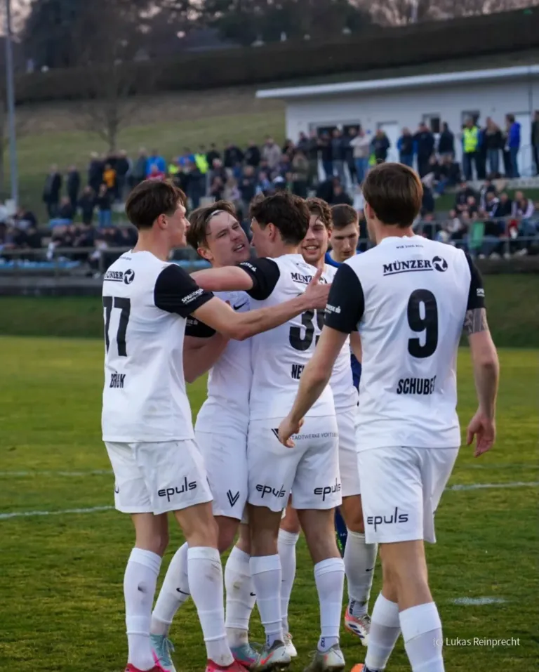 Fußballspieler in weißen Trikots feiern auf einem Rasenfeld, mit Trikots mit den Nummern 17, 3, 9 und 15, während eine Menge hinter einem Zaun zuschaut.