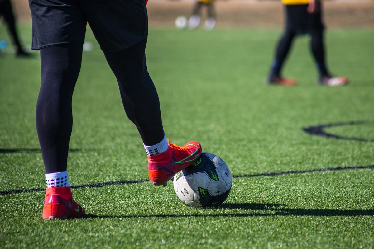 A soccer player's leg wearing red cleats is kicking a ball on a grassy field. Other players are blurred in the background.