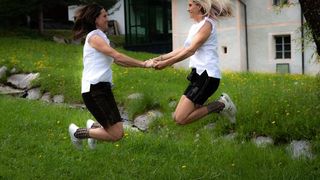 Two women jumping in a grassy yard, holding hands, with a house and greenery in the background.