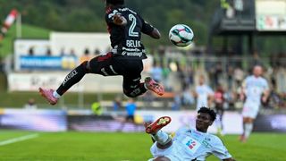 A soccer player in black jersey number 2 jumps to head the ball, while another player in white jersey looks on.