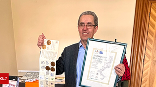 An older man in a gray suit holds a framed certificate and a booklet with coins, standing against a light-colored wall.