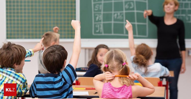 A classroom of children, with some raising their hands, sitting at their desks with a blackboard showing numbers in the background.
