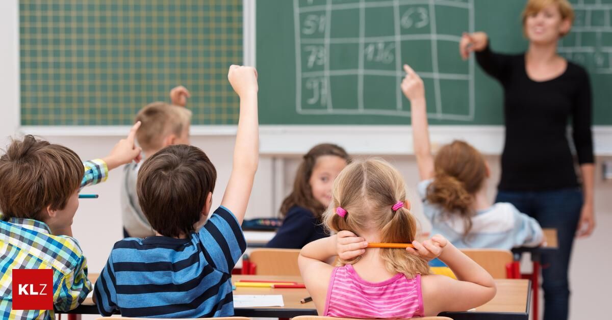 A classroom of children, with some raising their hands, sitting at their desks with a blackboard showing numbers in the background.