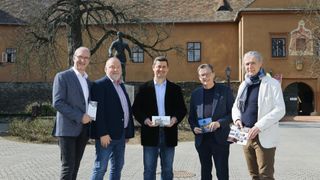 Five men are standing in front of a building with a statue of a man behind them. They are all smiling and holding brochures.