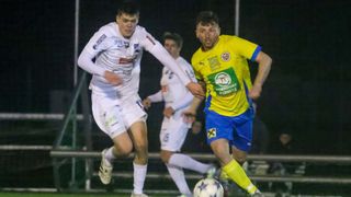 A soccer match with players in action, one in white and one in yellow with blue shorts, both chasing the ball. The background shows a blurred crowd.