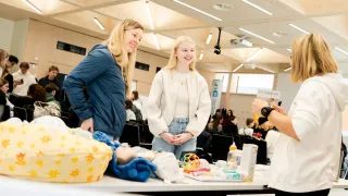 Two women are standing in a room filled with people, smiling and interacting. Behind them, a table is set up with various baby items, such as a doll, toys, and bottles.