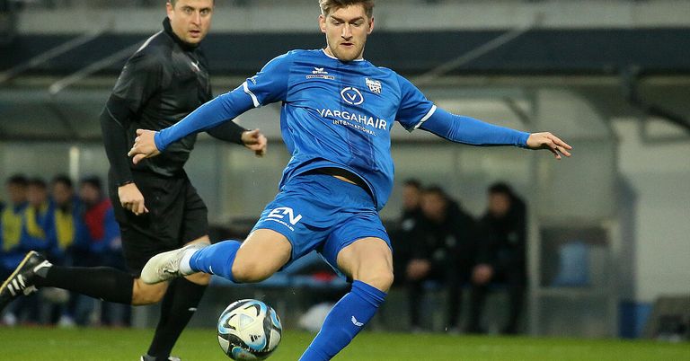 A soccer player in a blue jersey kicks the ball, while another player in black tries to block him. They are on a field with blurry people in the background.