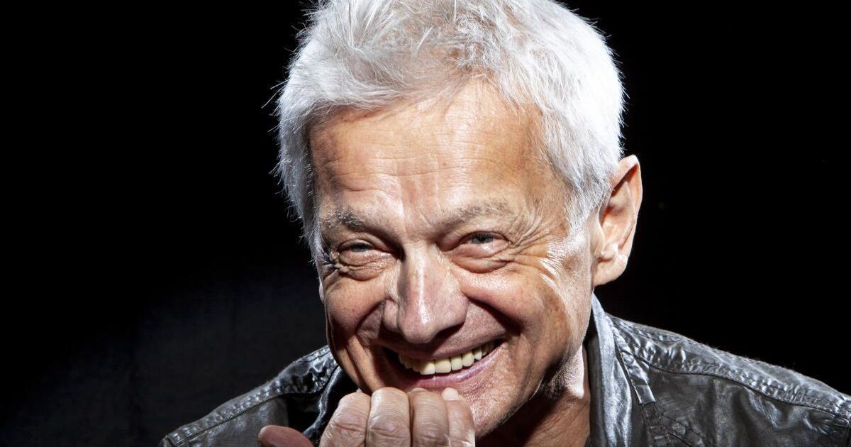 An older man with white hair smiles broadly, his hand resting on his chin. He wears a black leather jacket.