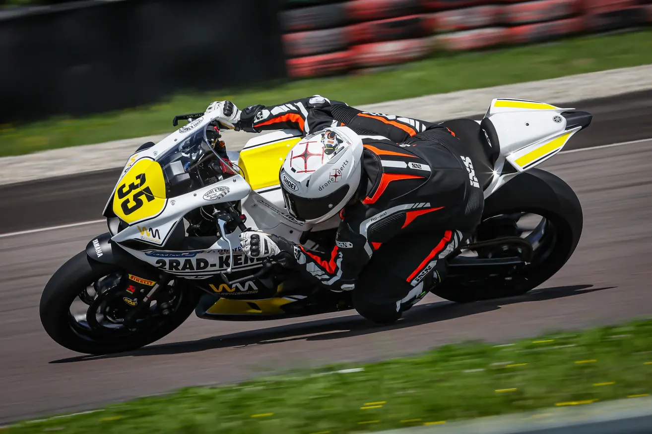 A motorcycle racer leans into a turn on a racetrack, wearing a helmet and racing suit. The motorcycle has a yellow number 53 on the side and is leaning to the side.
