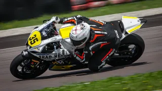 A motorcycle racer leans into a turn on a racetrack, wearing a helmet and racing suit. The motorcycle has a yellow number 53 on the side and is leaning to the side.
