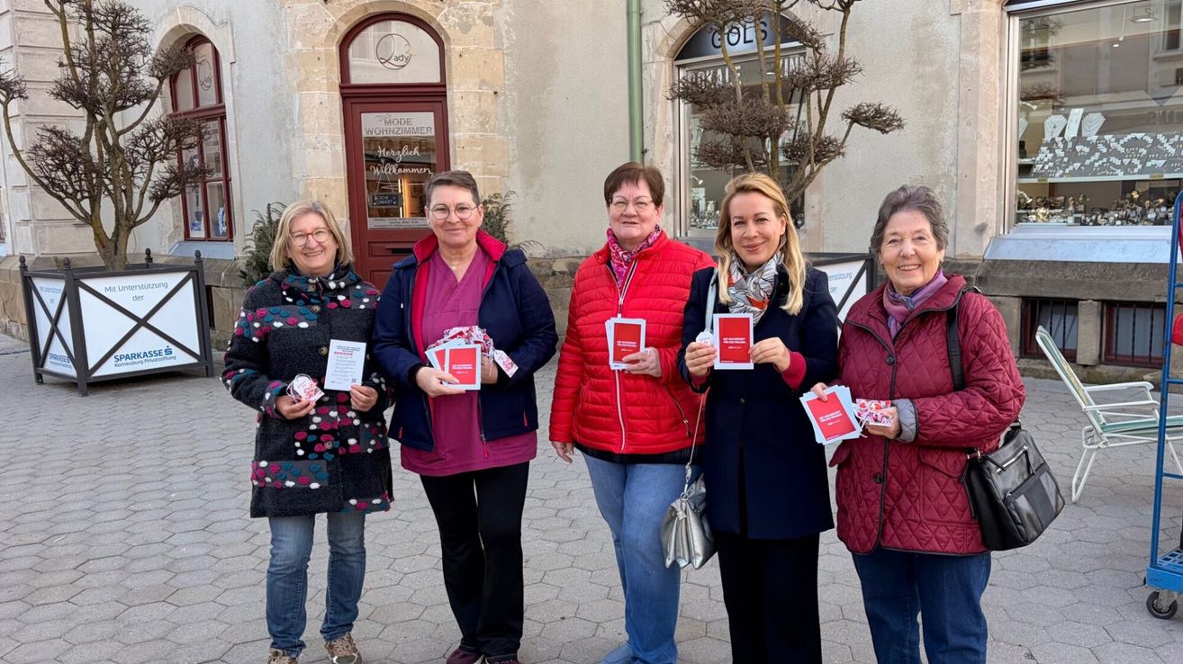 Five women stand in front of a building, holding red pamphlets. They are all smiling and seem to be posing for a photo. Behind them is a building with a sign that reads 'Mode Wohnzimmer'.