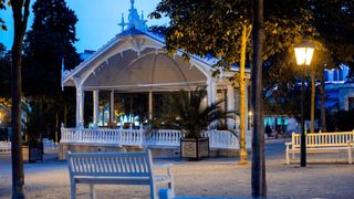 A beautifully lit gazebo with a white railing, a palm tree, and a bench in the foreground. It is surrounded by trees and has a fountain in the background.
