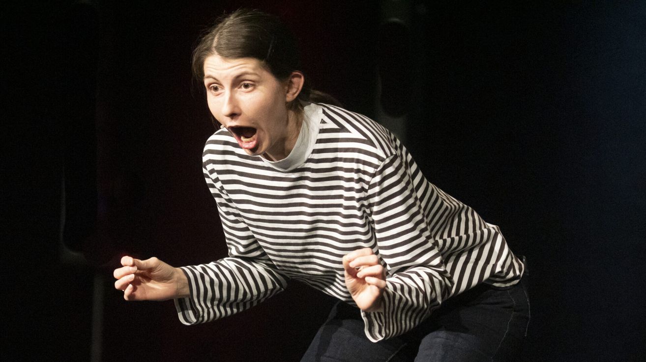 A woman in a black and white striped shirt appears to be shouting, her mouth open with a microphone near her lips. Her hands are extended outwards, and she is crouching slightly.