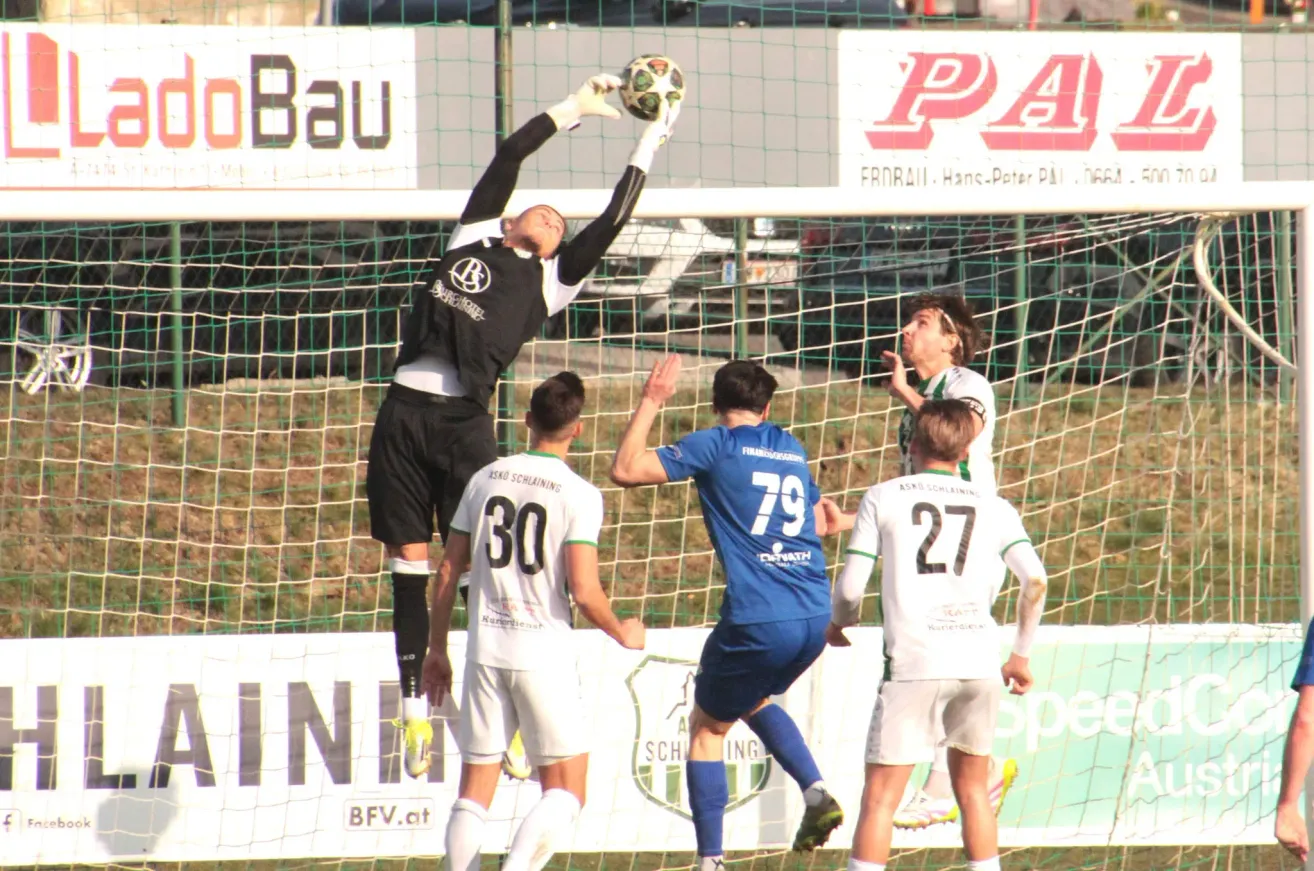 Soccer players in action during a match. A goalkeeper in black jumps to catch the ball, while others in blue and white jerseys attempt to score.
