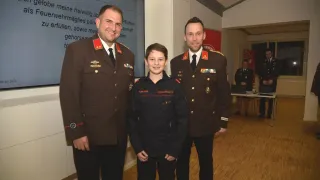 Three men in military uniforms stand together smiling for a photo. The middle man wears a cadet uniform. Behind them is a large screen with text.
