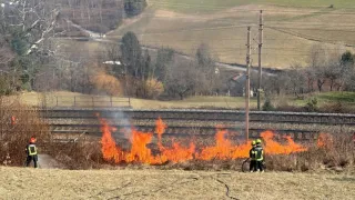 Two firefighters are extinguishing a fire near a railway track. The fire is surrounded by dry grass and bushes. Power lines and a pole are in the background.