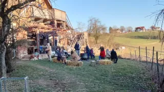 An outdoor gathering with people sitting on haystacks around a wooden house. Some individuals are standing near the house and others are sitting on benches. Trees and a house are in the background.