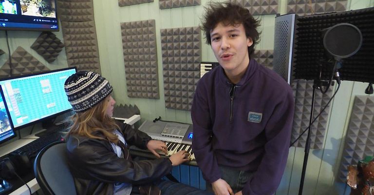 Two people in a music studio, a girl playing the piano and a boy standing next to her.