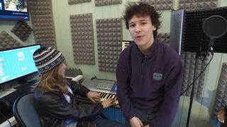 Two people in a music studio, a girl playing the piano and a boy standing next to her.
