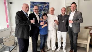 A group of five people are posing for a photo, holding wine glasses. They are smiling and appear to be at a party. A woman in the middle holds two glasses. Behind them is a couch and a painting.