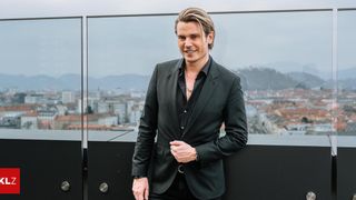 A man in a black suit, with blonde hair, stands on a rooftop balcony with a cityscape view, smiling and posing.