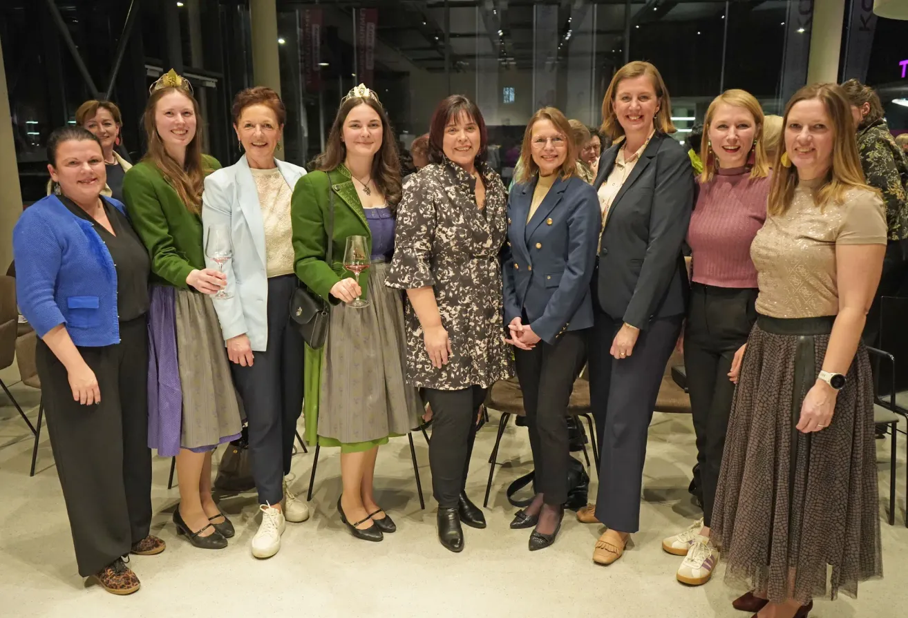A group of women stand together indoors, smiling for a photo. Some hold wine glasses. They wear various colored blazers and dresses. One woman wears a crown, and others have accessories. They stand on a light-colored floor.