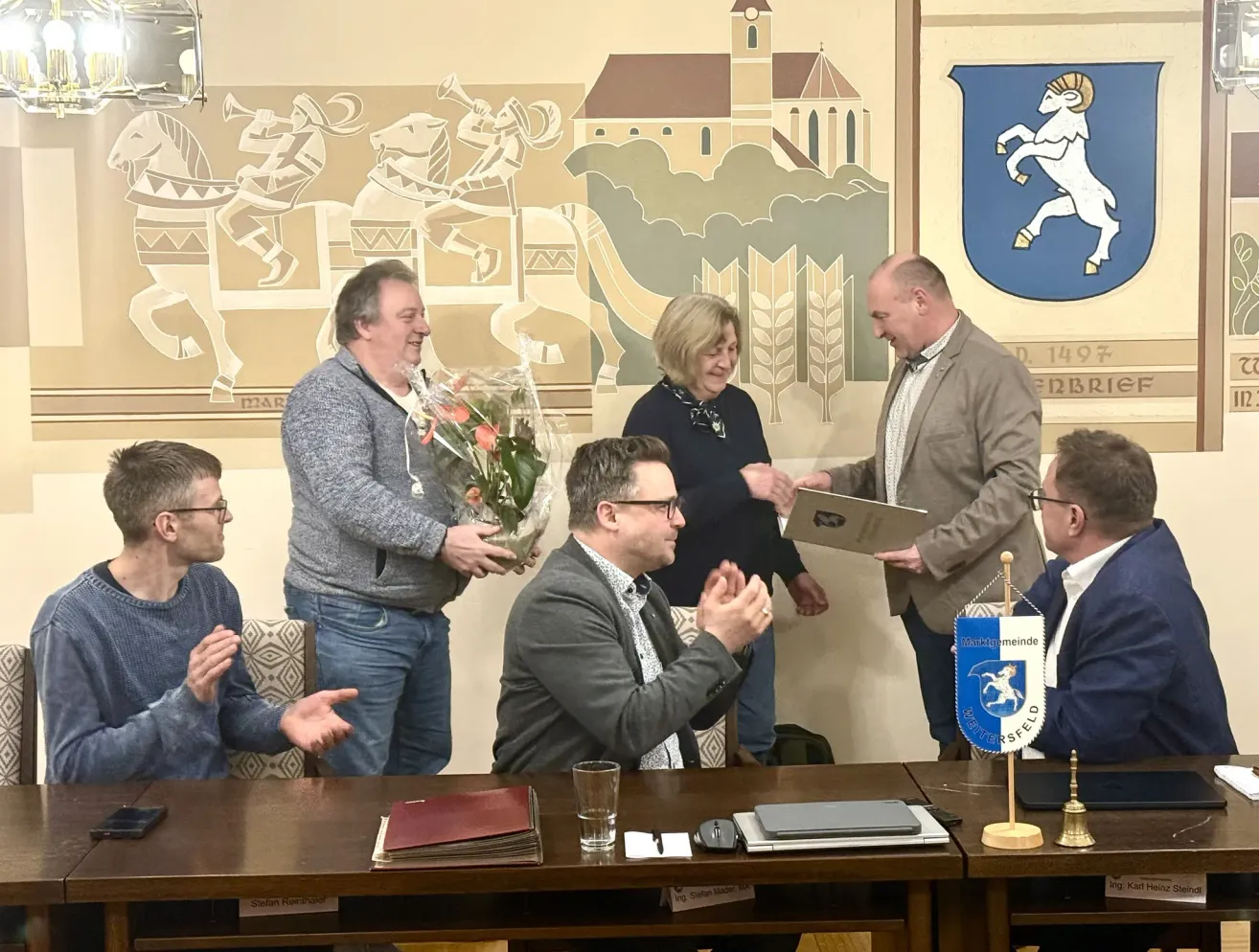 Eine Gruppe von Menschen versammelt sich lächelnd um einen Tisch. Ein Mann hält einen Blumenstrauß und ein Zertifikat, während ein anderer es präsentiert. Sie sind von verschiedenen Gegenständen wie Büchern, einem Glas und einer Glocke umgeben. Hinter ihnen befindet sich eine Wand mit einem Wandbild und einem Wappen.