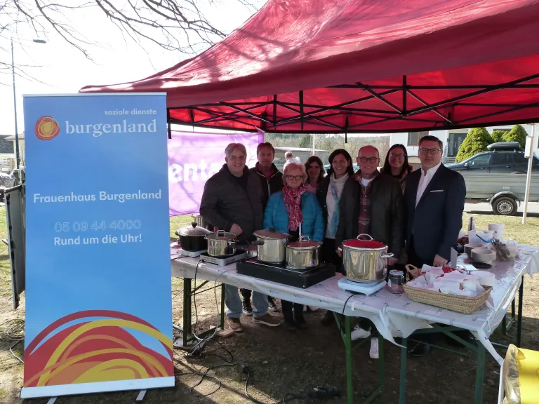 Mehrere Personen versammeln sich unter einem roten Zelt und lächeln für ein Foto. Ein Tisch mit Töpfen und Pfannen ist aufgebaut, mit einem blauen Banner und einem Banner mit der Aufschrift 'soziale dienste' in der Nähe.