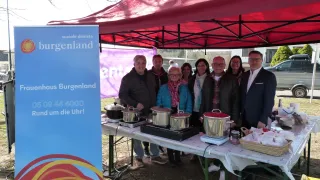 Several people gather under a red tent, smiling for a photo. A table with pots and pans is set up, with a blue banner and a banner reading 'soziale dienste' nearby.