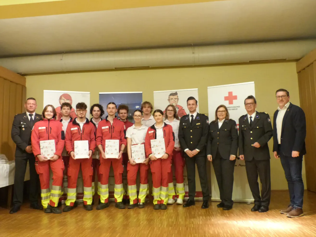 A group of individuals, some in red jumpsuits and others in black uniforms, pose for a photo. They hold certificates and stand in front of banners with a red cross.