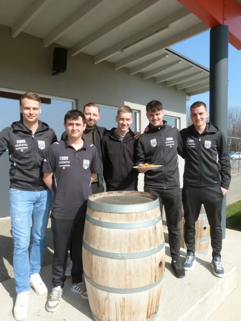 Six men in matching black jackets pose for a photo in front of a building with a wooden barrel. Each man holds a plate of food, and they are smiling.